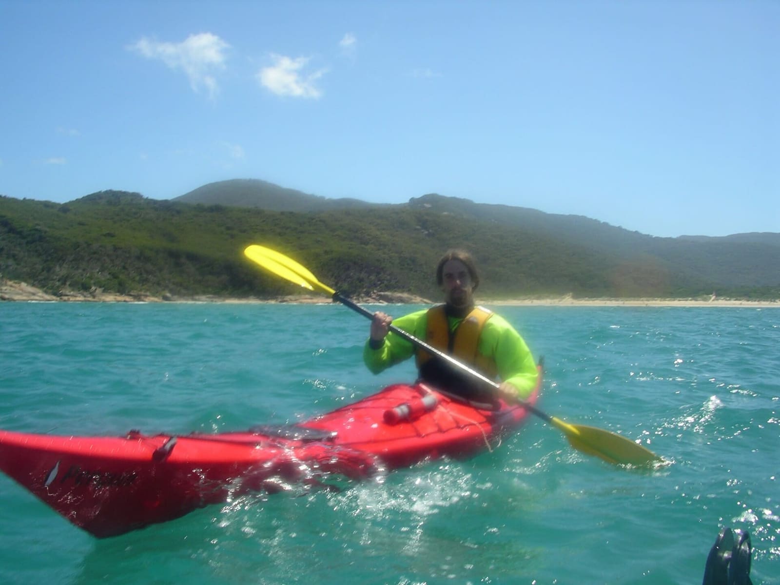 A sea kayaker in a red kayak on turquoise water with forested hills behind
